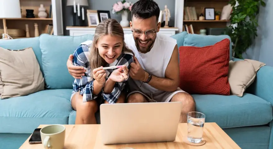 Szczęśliwa para z testem ciążowym na ekranie laptopa. Czy da się zaplanować płeć dziecka? Ich radość sugeruje, że odkryli coś ekscytującego.