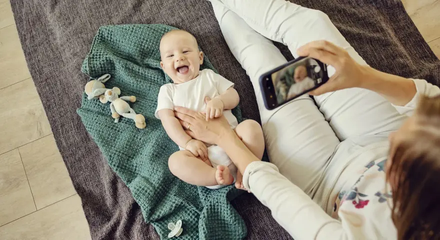 Uśmiechnięte niemowlę na kocyku, mama robi zdjęcie telefonem. To jeden ze sposobów, jak wspierać rozwój niemowlaka – dokumentując jego radość.