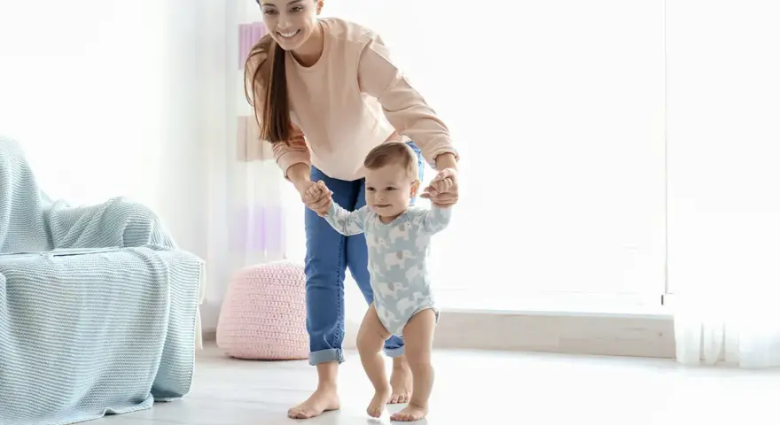 Uśmiechnięta mama pomaga dziecku stawiać pierwsze kroki. Pokazuje, jak trzymać dziecko przy nauce chodzenia, wspierając je za rączki.