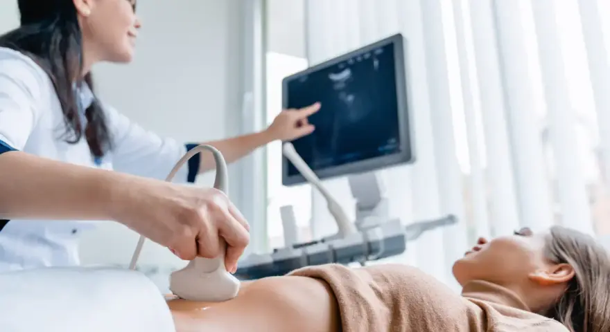Lekarka wykonuje badania prenatalne 1 trymestr, przykładając głowicę USG do brzucha pacjentki. Na ekranie widoczny obraz.