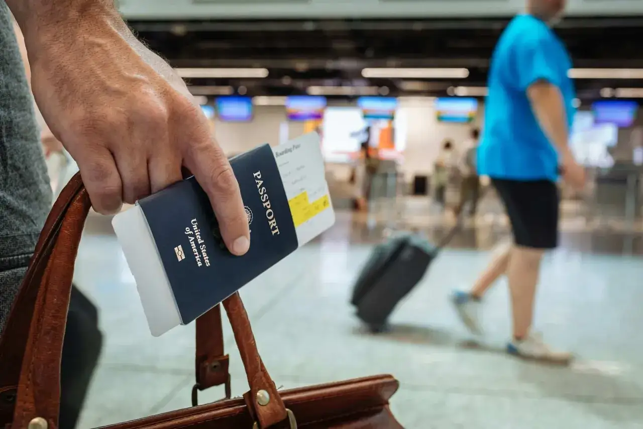 Ręka trzyma paszport i kartę pokładową, gotowa do kontroli paszportowej na lotnisku. W tle widać innych podróżnych.