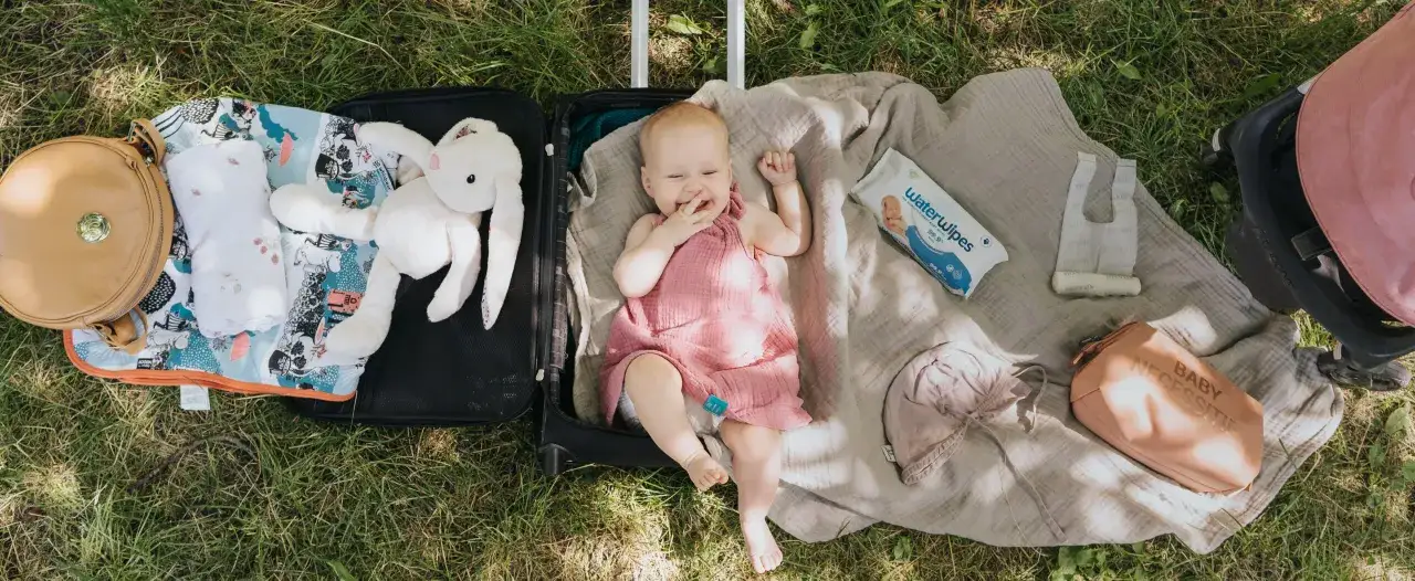 Uśmiechnięte niemowlę w walizce, gotowe na podr&oacute;ż z niemowlakiem. Obok pluszowy kr&oacute;liczek, kocyk, chusteczki i kosmetyczka.