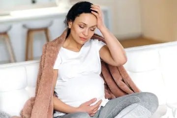 Kobieta w ciąży z bólem głowy, okryta kocem, szuka informacji co na przeziębienie w ciąży.
