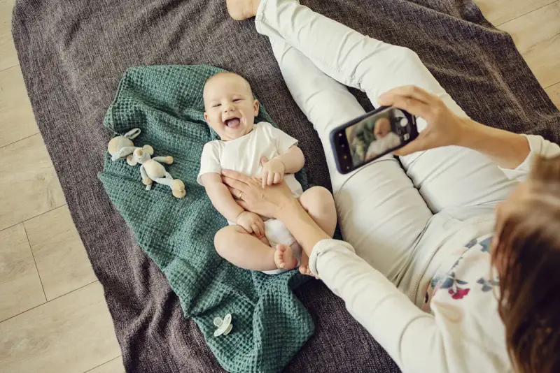 Uśmiechnięte niemowlę na kocyku, mama robi zdjęcie telefonem. To jeden ze sposobów, jak wspierać rozwój niemowlaka – dokumentując jego radość.