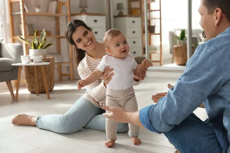 Rodzice zachęcają dziecko do pierwszych kroków. Uśmiechnięta mama i tata z otwartymi ramionami motywują malucha do chodzenia.